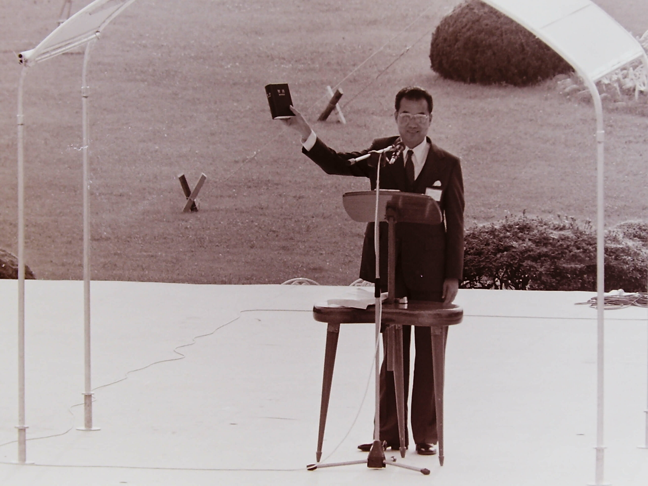 1982 調布 新世界訳聖書の発表 an old photo with a man standing behind a lectern holding a Bible in a hand raised in the air