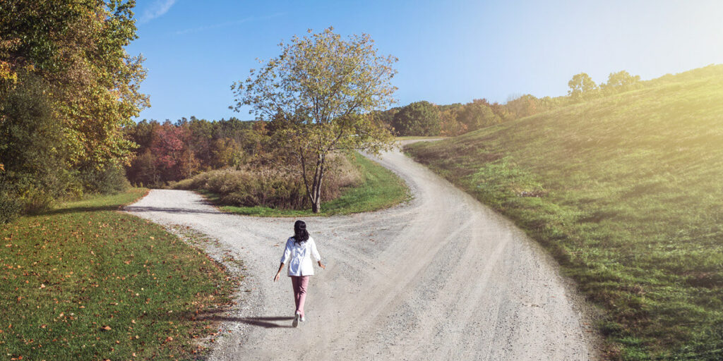 a woman walking along a path on a bright sunny day facing a fork in the road with two diverging paths