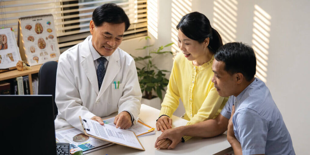 a doctor in his office sitting and talking with a couple while showing a document on a clipboard